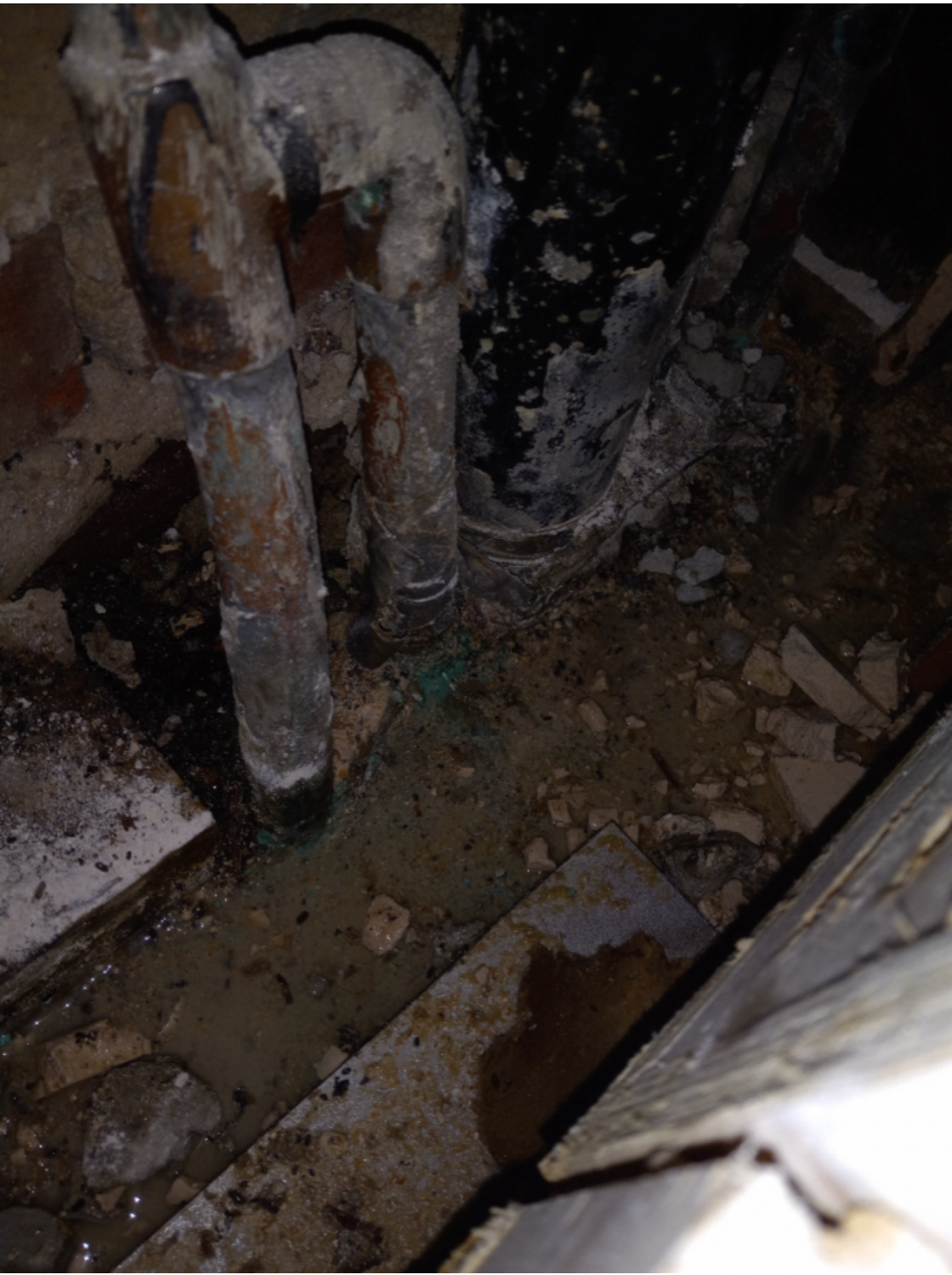 Rusty pipes and debris in a dark, damaged corner with a wet, dirty floor