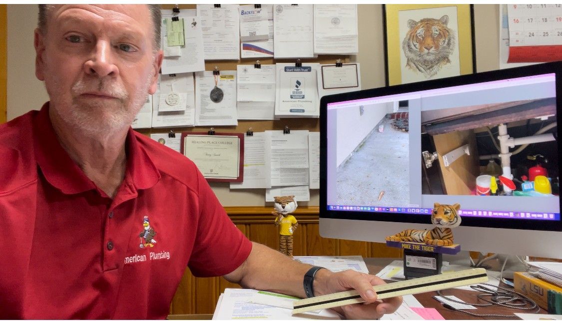 Man in red shirt with a computer screen showing a room; he holds a ruler, sits at a desk with items.