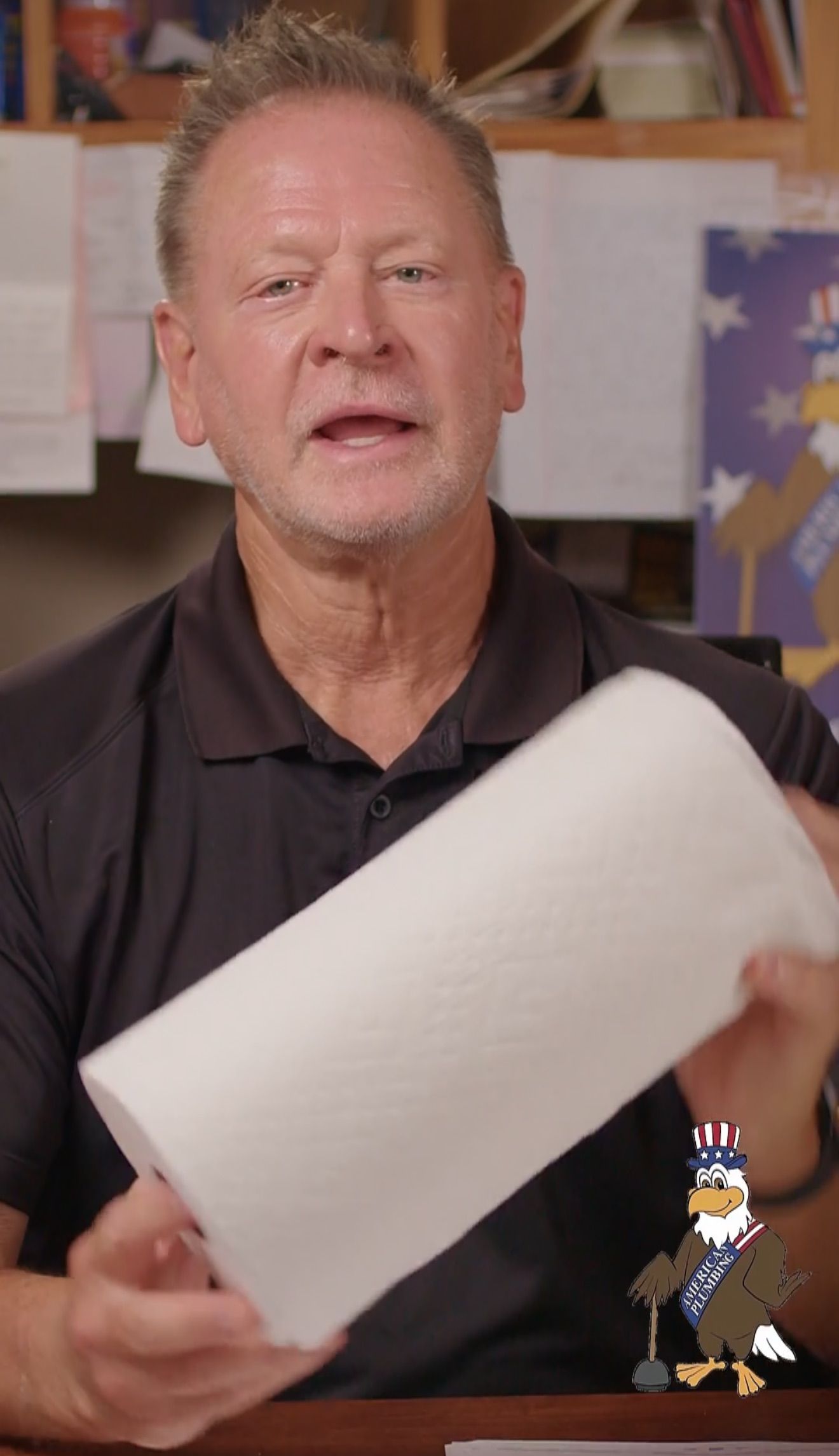Man holding a large white rolled sheet in an office, with a bulletin board behind him.
