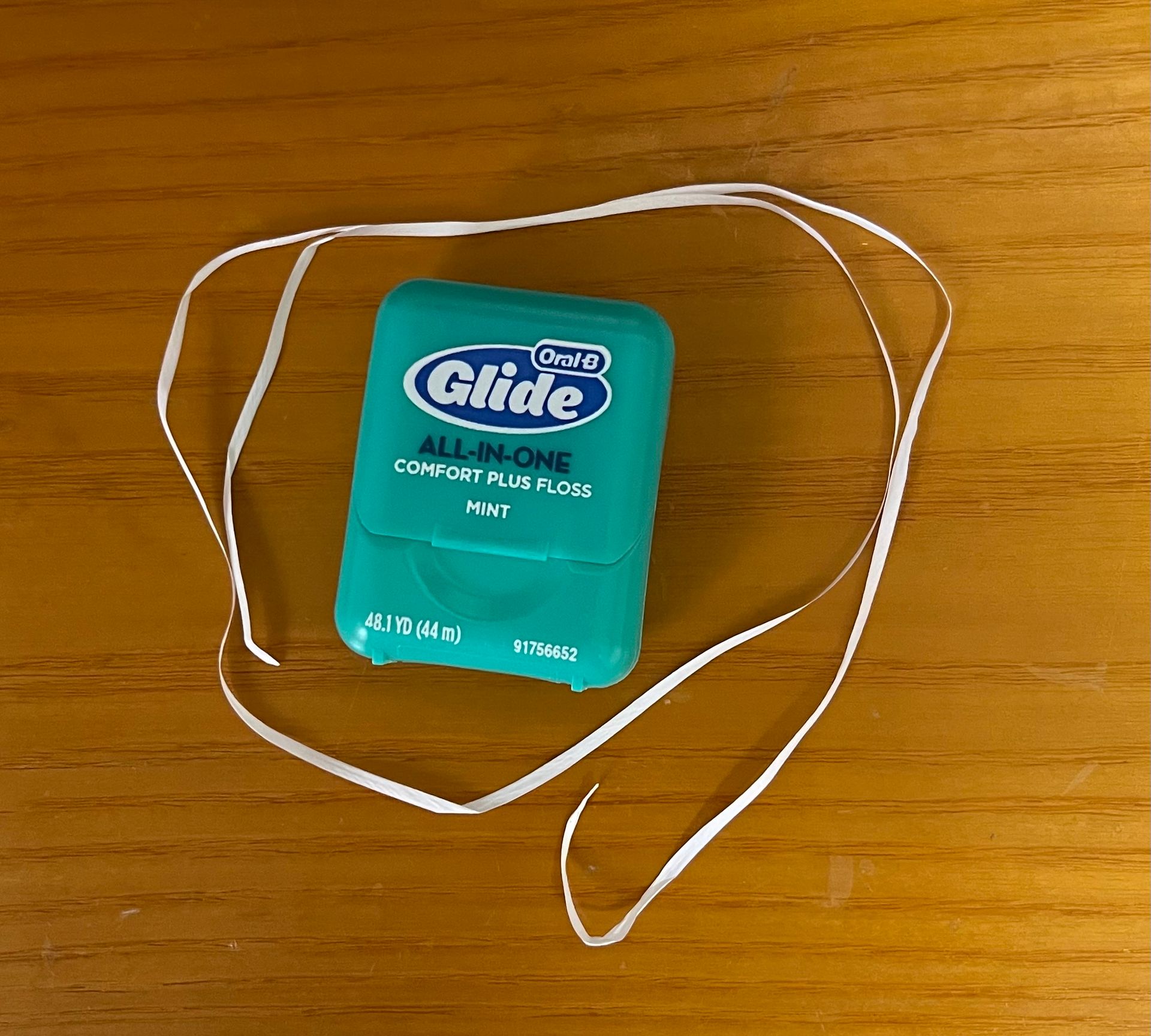 White dental floss on a wooden surface beside a turquoise Glide container.