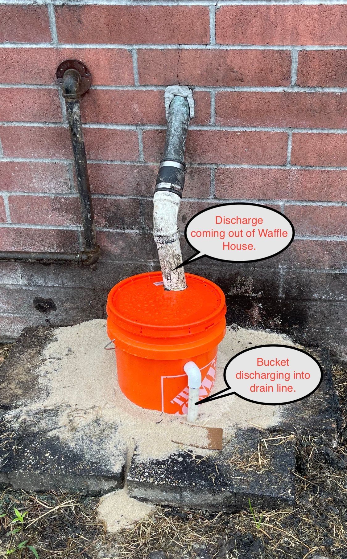 Orange utility bucket with exposed pipes against a brick wall and ground damage nearby.