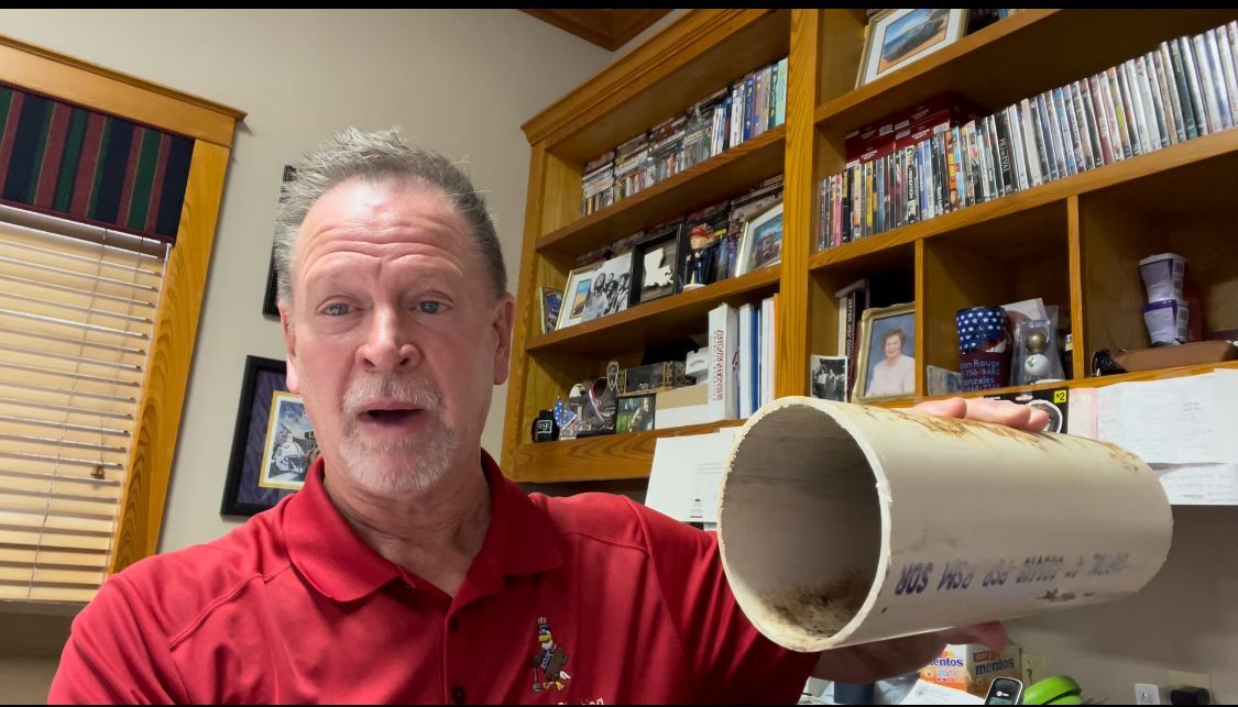 Man in red shirt holding a white PVC pipe, talking. Bookshelves in background.
