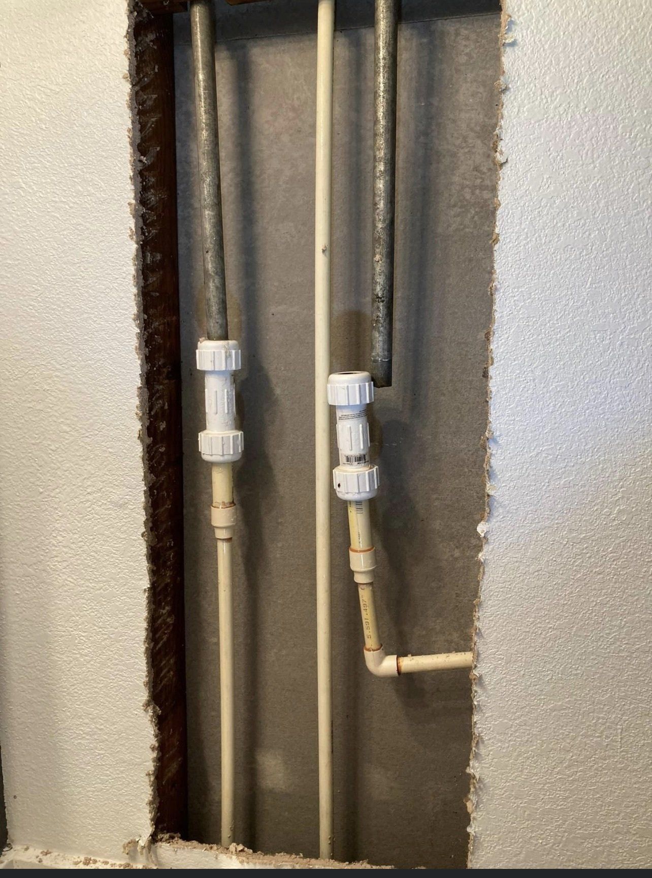 Pipes in a wall cavity, with white plastic fittings connecting metal and plastic pipes. Foam insulation surrounds the cavity.