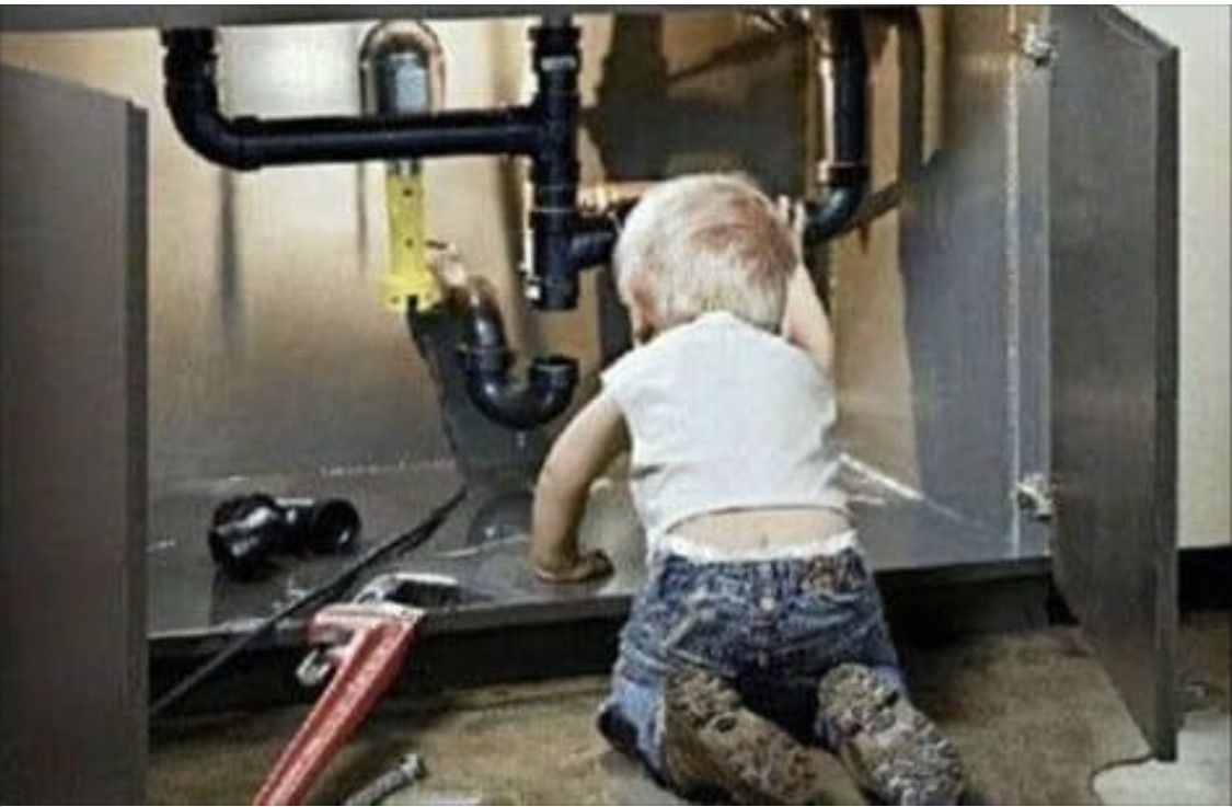 Child kneeling under a sink, reaching toward plumbing pipes with tools scattered nearby.