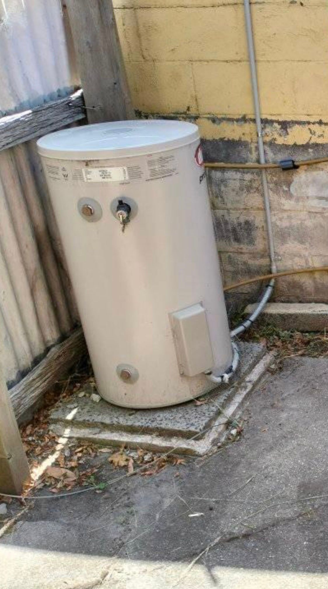 Outdoor beige cylindrical water heater beside a wall in a narrow concrete yard