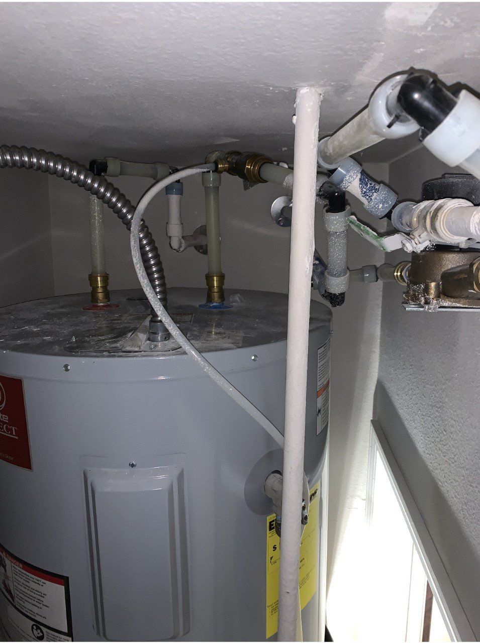 Water heater in a cramped utility space with exposed pipes and a white pole in front.