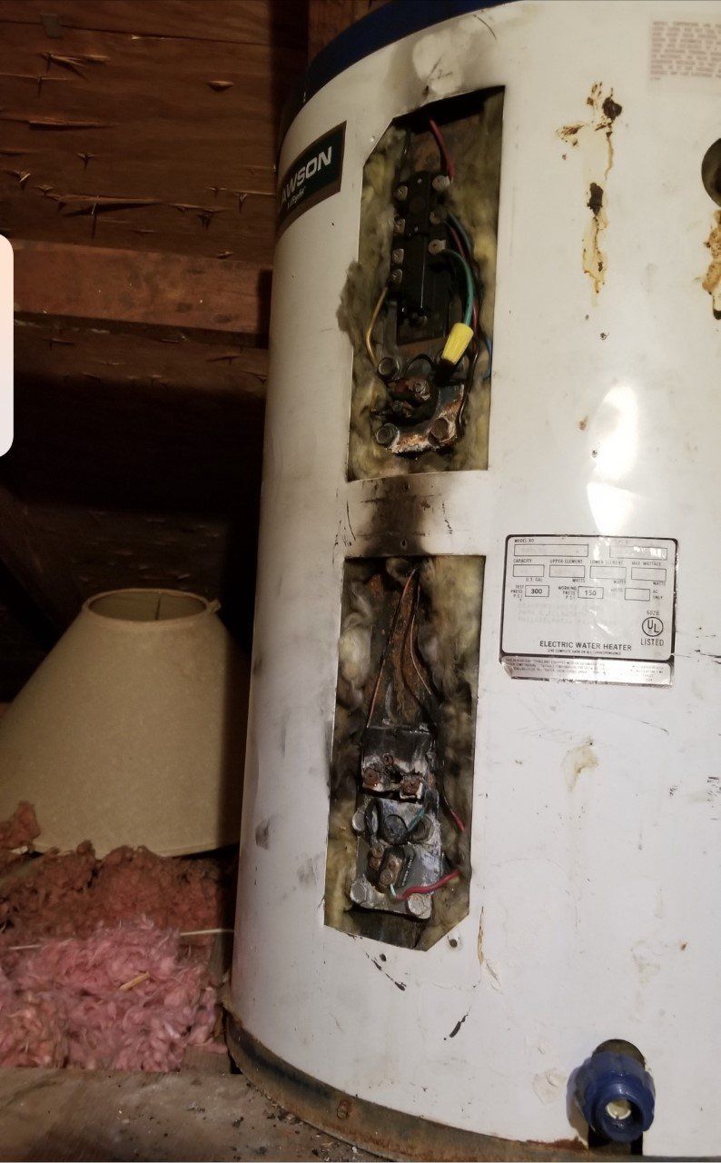 Damaged electric water heater with charred sections and exposed wiring in an attic.