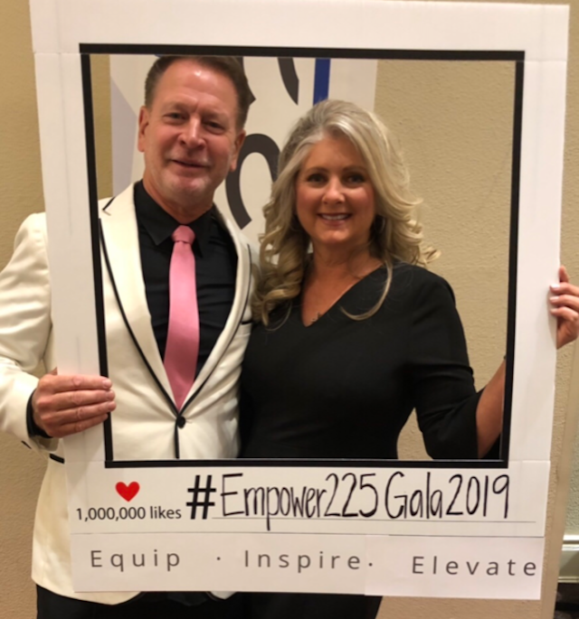 Man and woman framed by a photo prop, smiling. White jacket, pink tie. Black dress. 