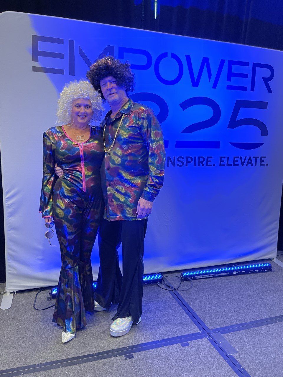 Two people posing in front of an “EMPOWER 2025” event backdrop under blue lighting