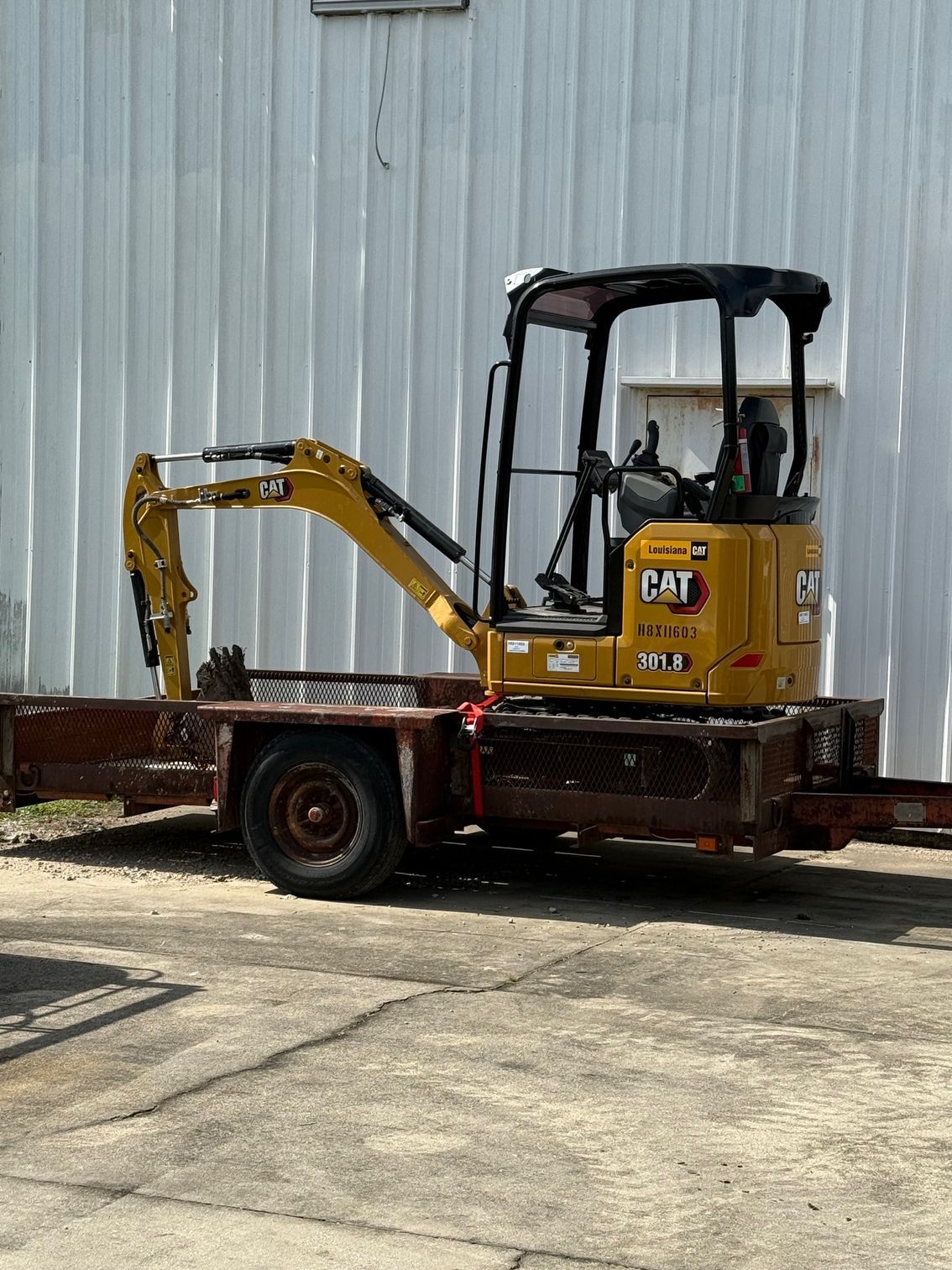 Yellow Caterpillar mini excavator on a brown flatbed trailer, parked in front of a white building.