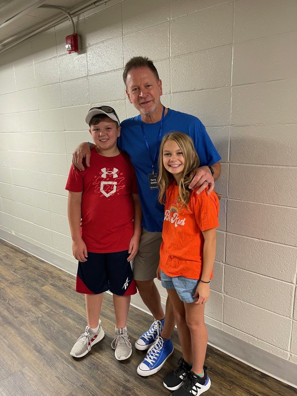 Man with arms around two smiling children in a hallway; man wears blue shirt, children wear red/orange shirts.