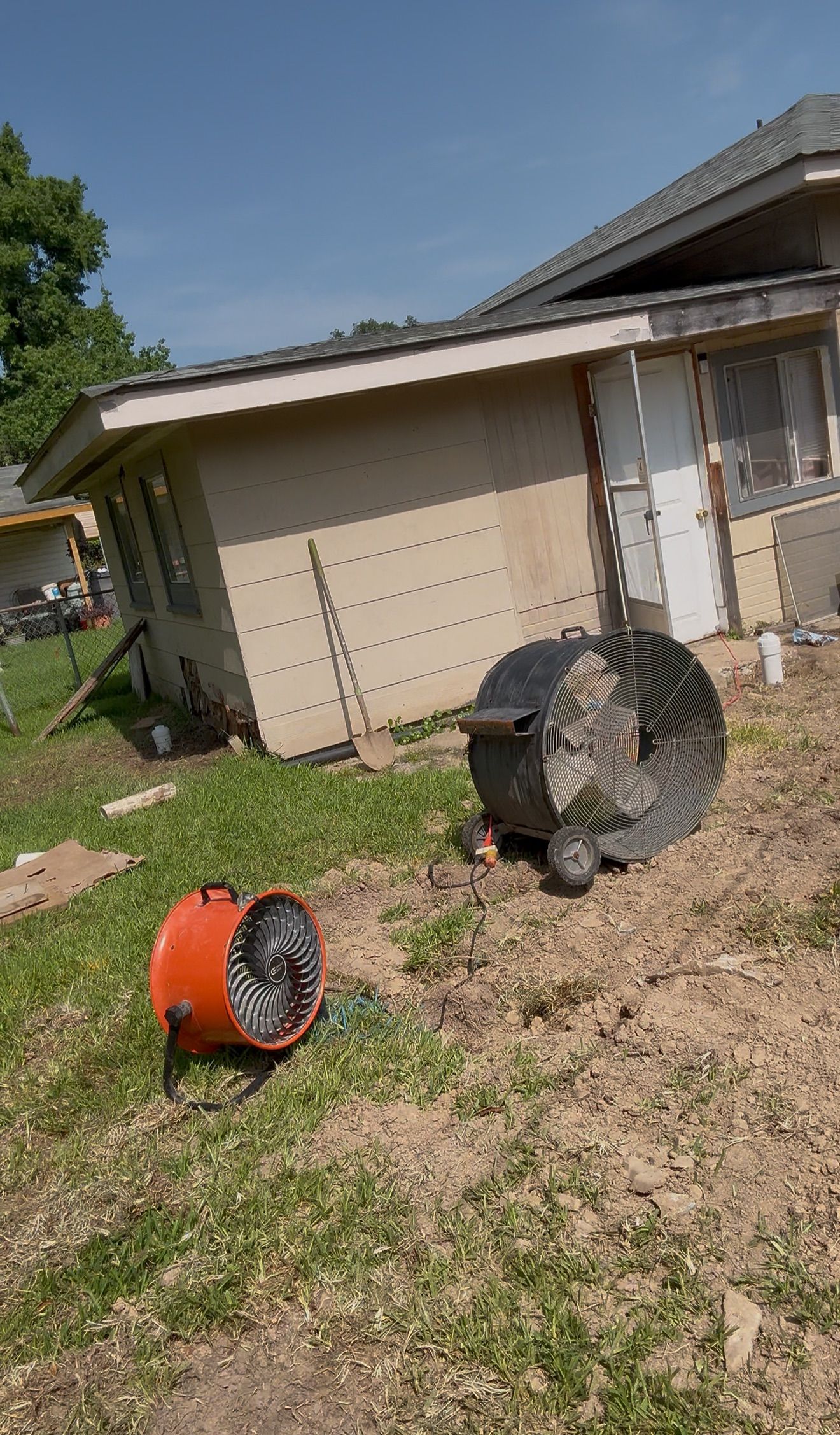 Two portable fans, one orange and one black, sit on a grassy yard beside a beige house.