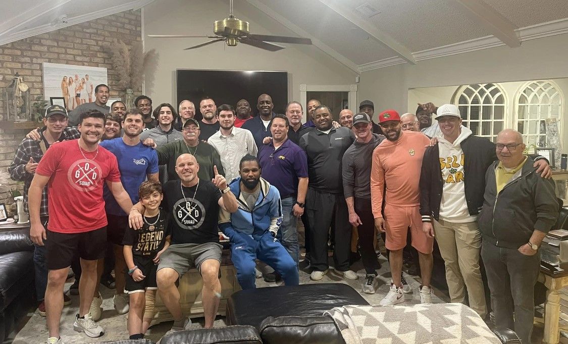 Large group posing together in a living room, smiling and standing around a couch under a ceiling fan.