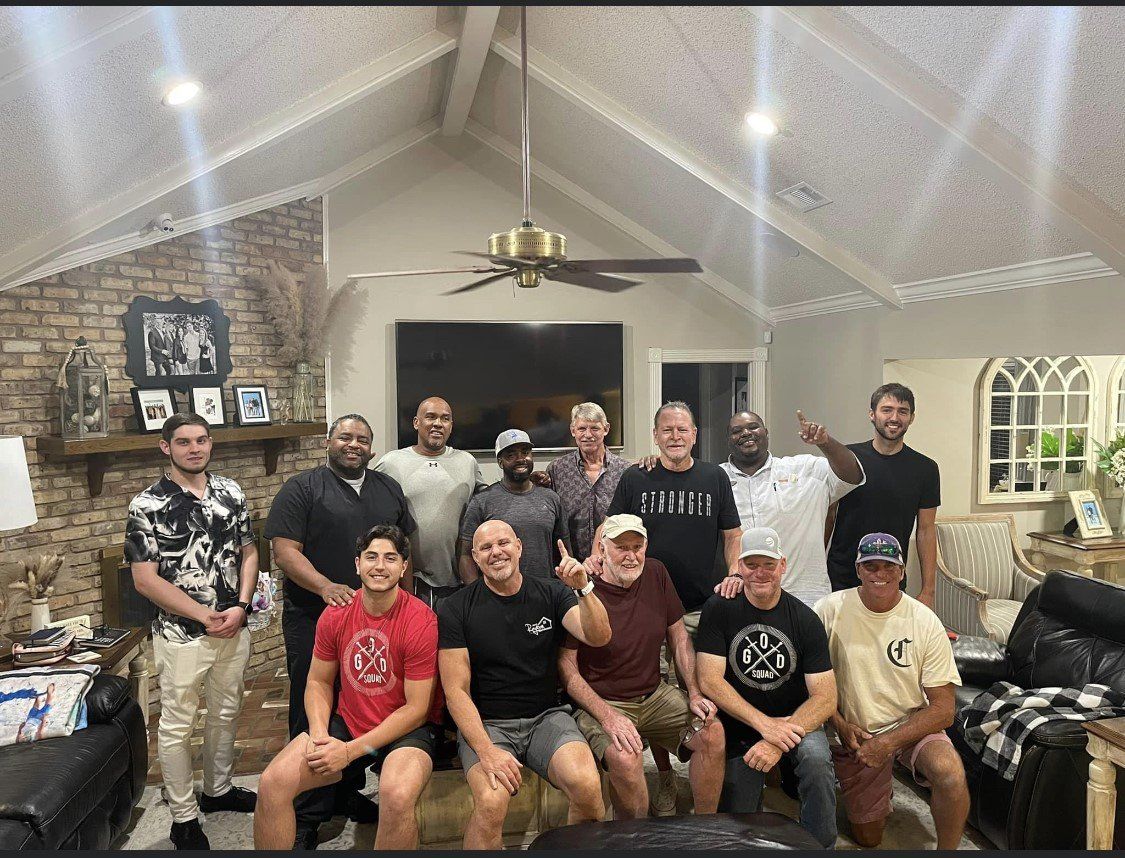Group of men smiling and posing indoors, some seated, in a living room with brick fireplace and vaulted ceiling.