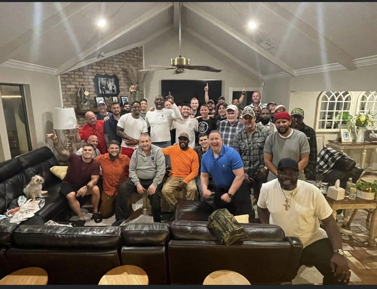 Large group posing together in a living room with sofas, lamps, and a chandelier, smiling at the camera.