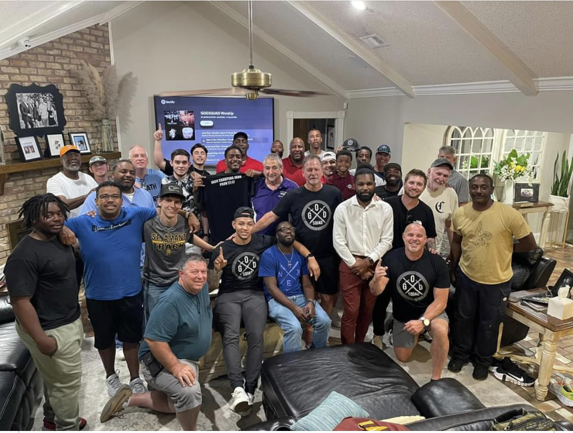 Group of men smiling indoors, gathered around a TV. Some pose, others cheer. Bright room, casual attire.