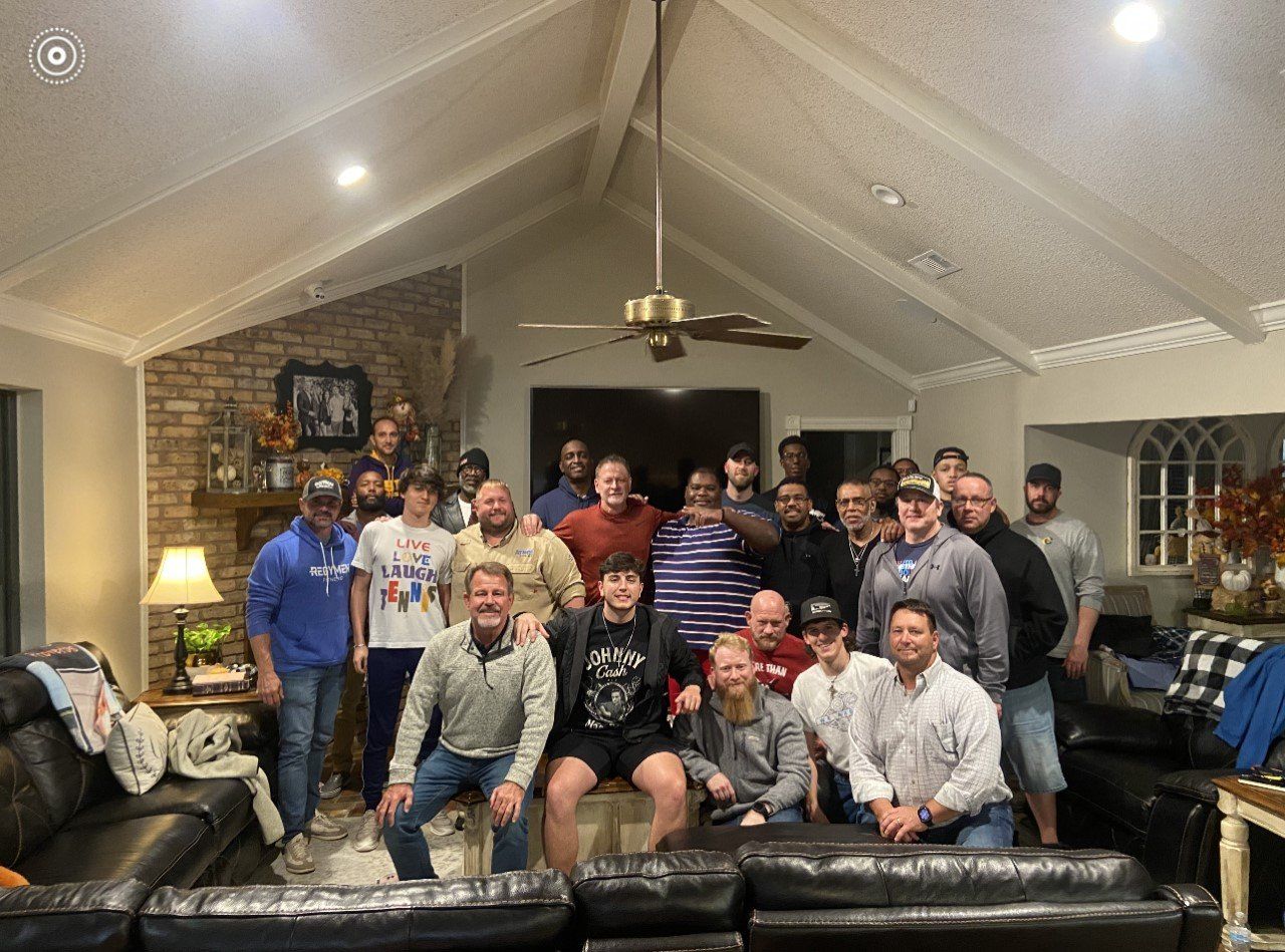 Group of men in a living room, posing for a photo. Some are sitting on couches, others standing, smiling.