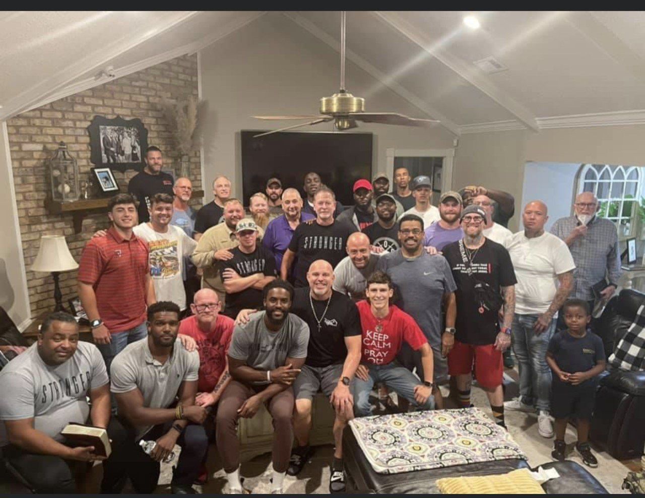 Large group of men posing inside a home. Brick wall, ceiling fan, and several smiles are visible.