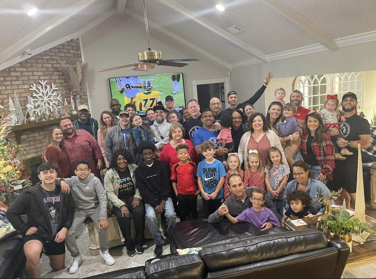 Large group posing in a living room, smiling in front of a colorful banner and fireplace.