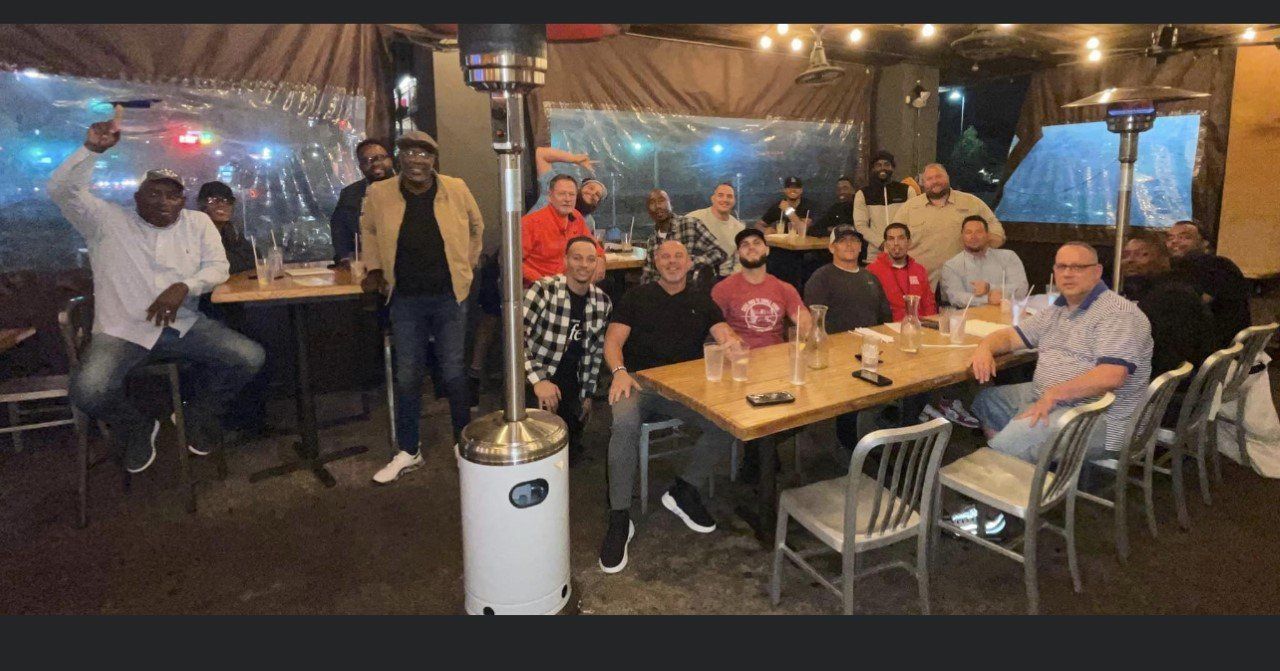 Group of men at outdoor restaurant. Some sitting, some standing, smiling. Nighttime.