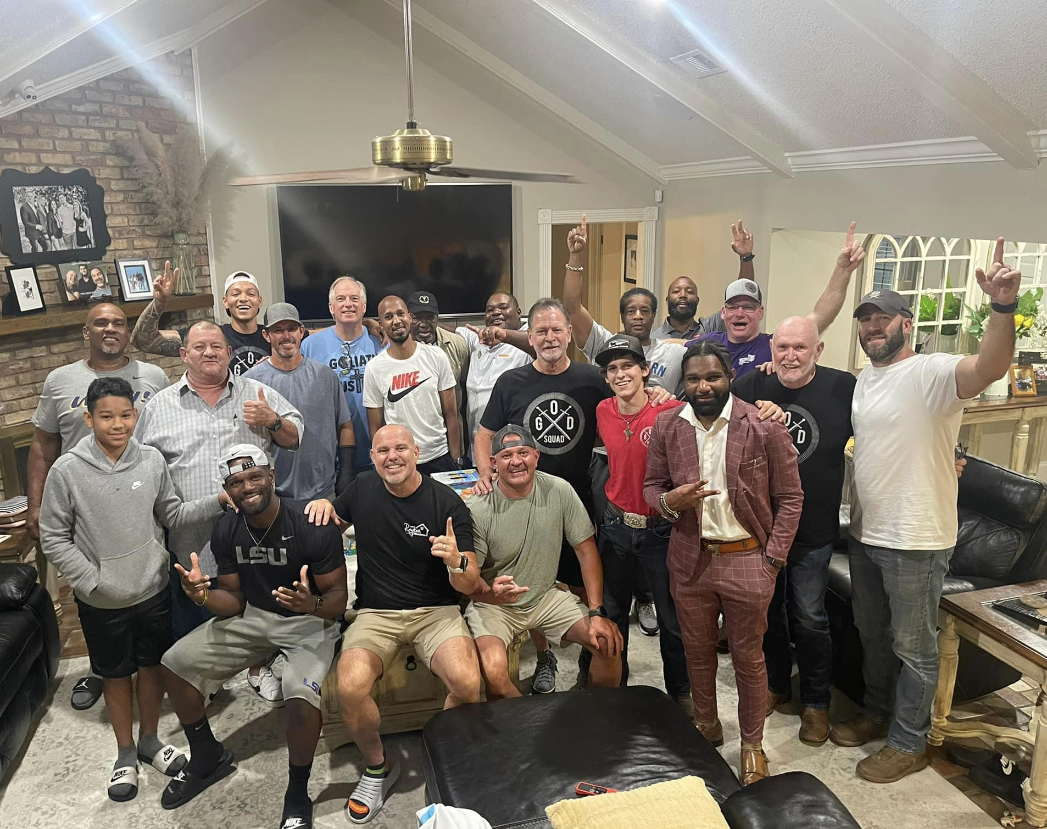 Group of men celebrating indoors; some pose, raise hands, and smile. A living room setting.