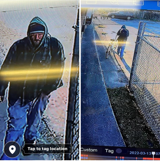 Person in dark hooded jacket and blue pants walking on a snowy sidewalk beside a chain-link fence.