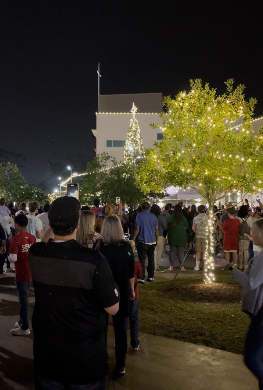 Crowd gathered outdoors at night near a lit building and trees, with people walking along a pathway