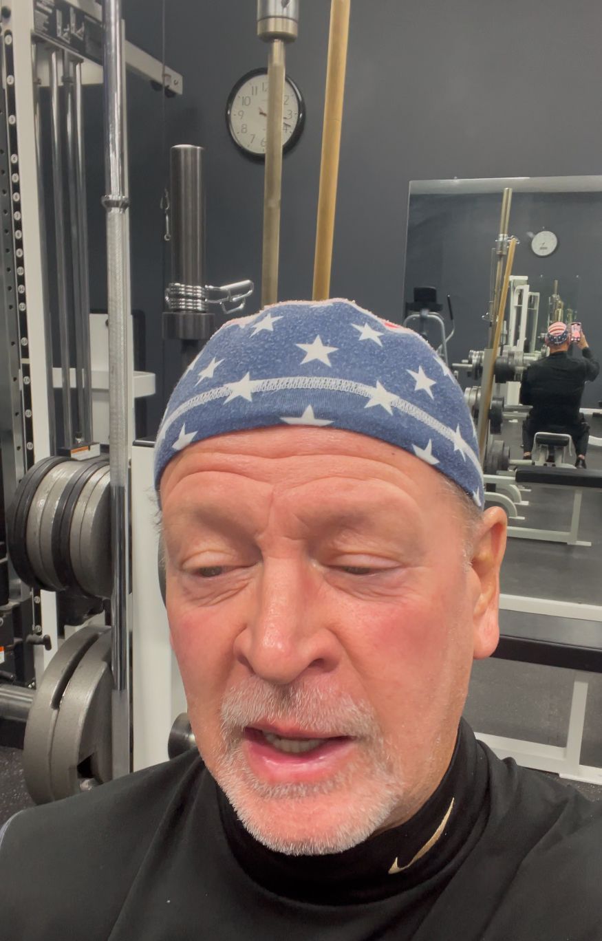 Man in a gym wearing a blue bandana; speaking. Other gym equipment and a mirror in the background.