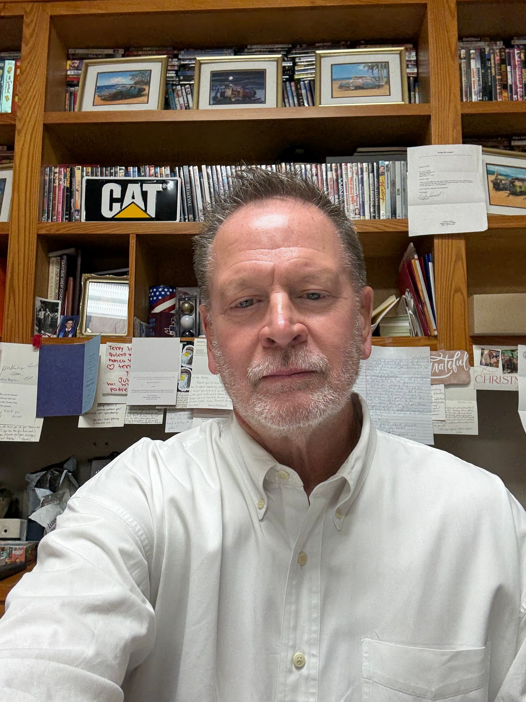 Man with a gray beard in a white shirt in front of a bookshelf with a 
