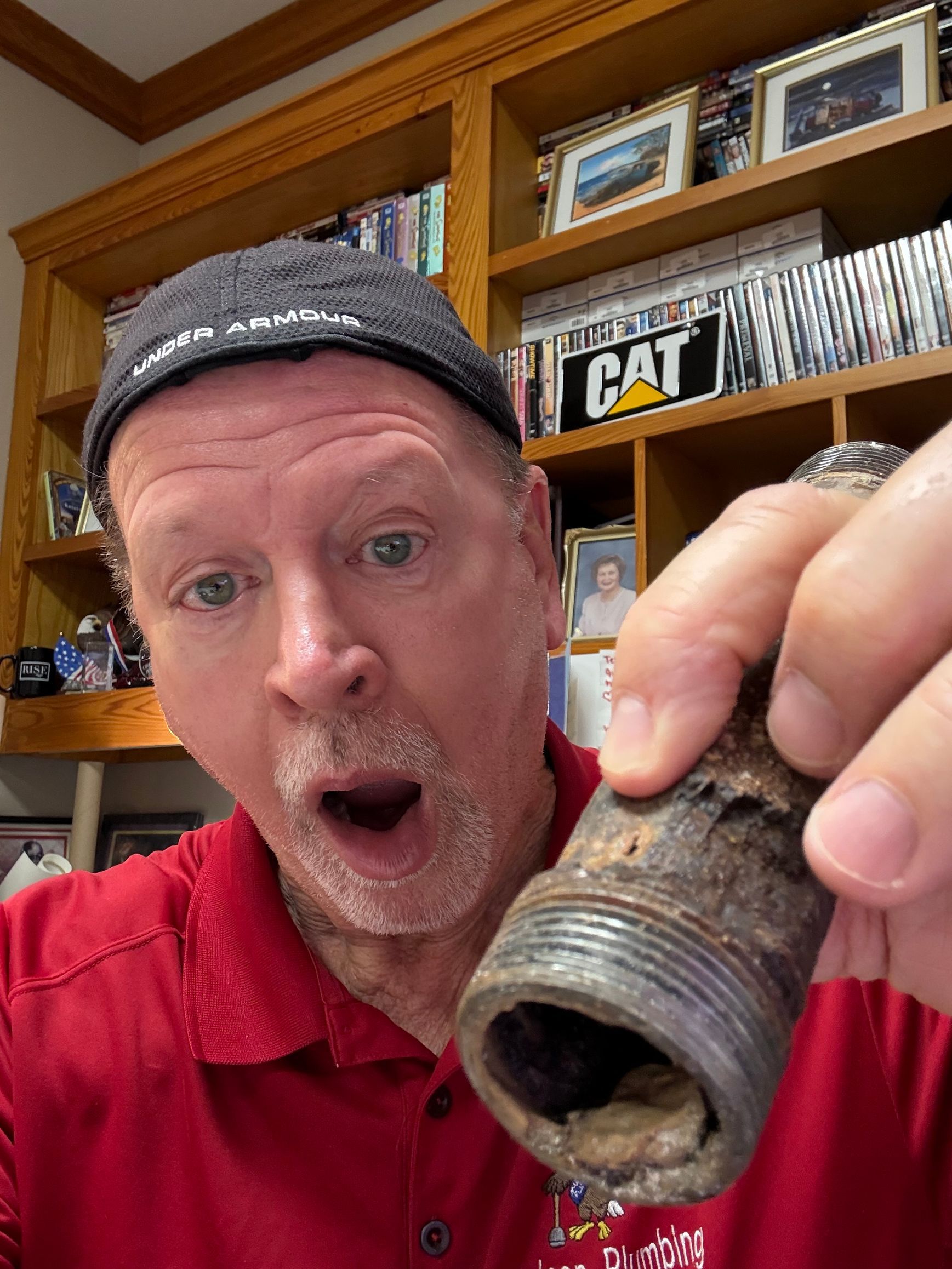 Man in red shirt holding up a rusty pipe fitting in a workshop, surprised expression.