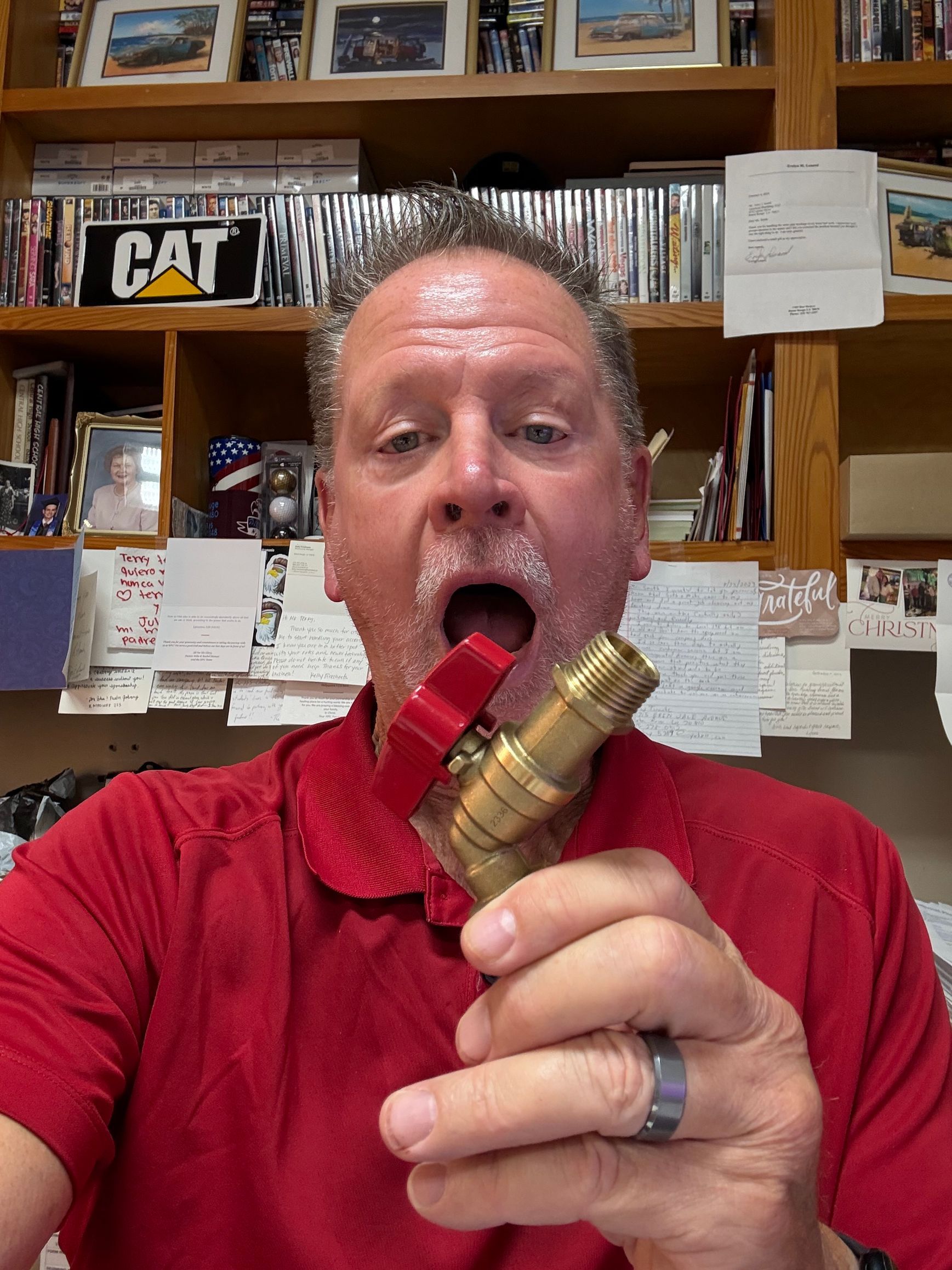 Man in a red shirt holding a brass fitting up to his open mouth in a workshop or store aisle