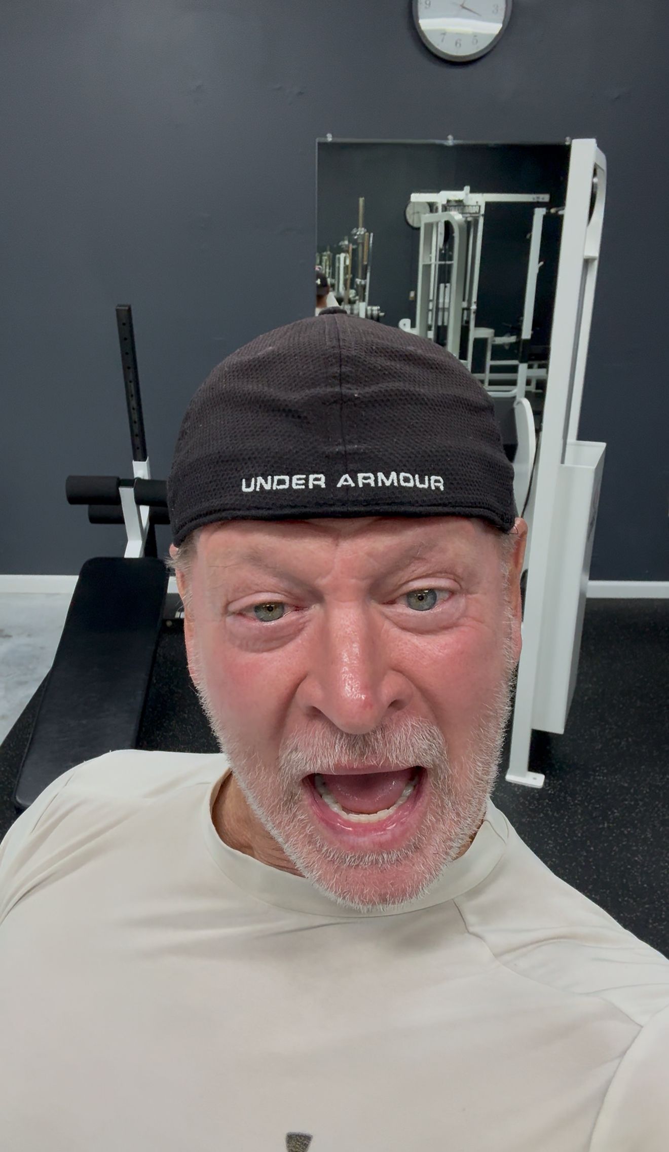 Man posing in a gym, wearing a black headband and light shirt, with workout equipment behind him.