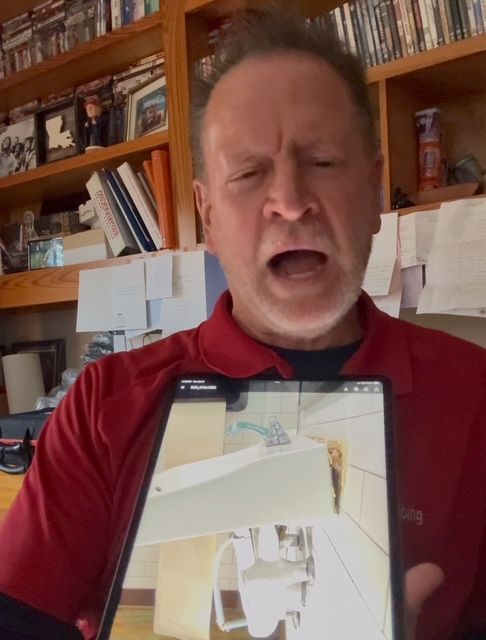 Man in red shirt holds tablet, looking surprised while showing a bathroom scene.