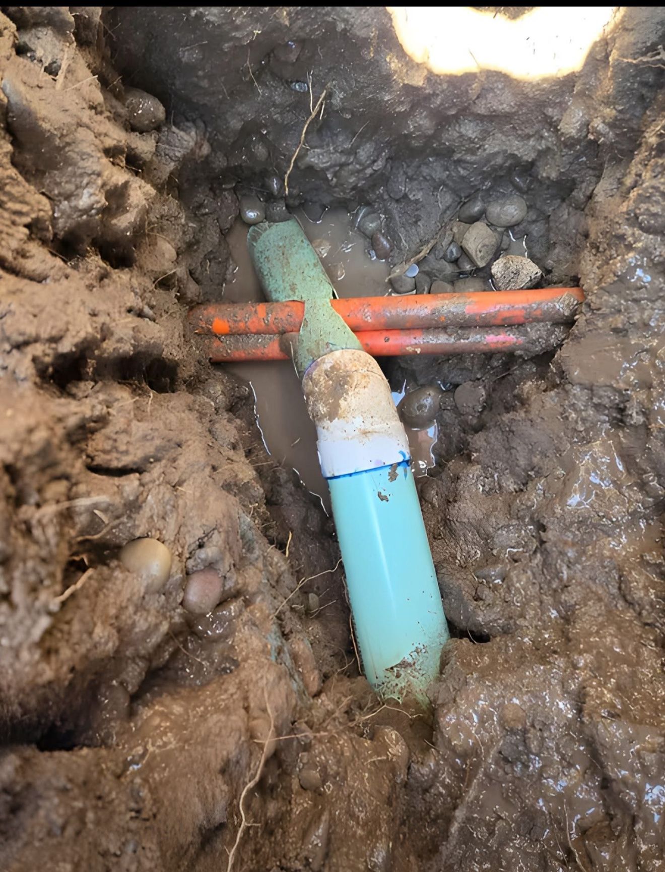 Trench with exposed green and orange pipes in muddy soil, showing a plumbing repair.