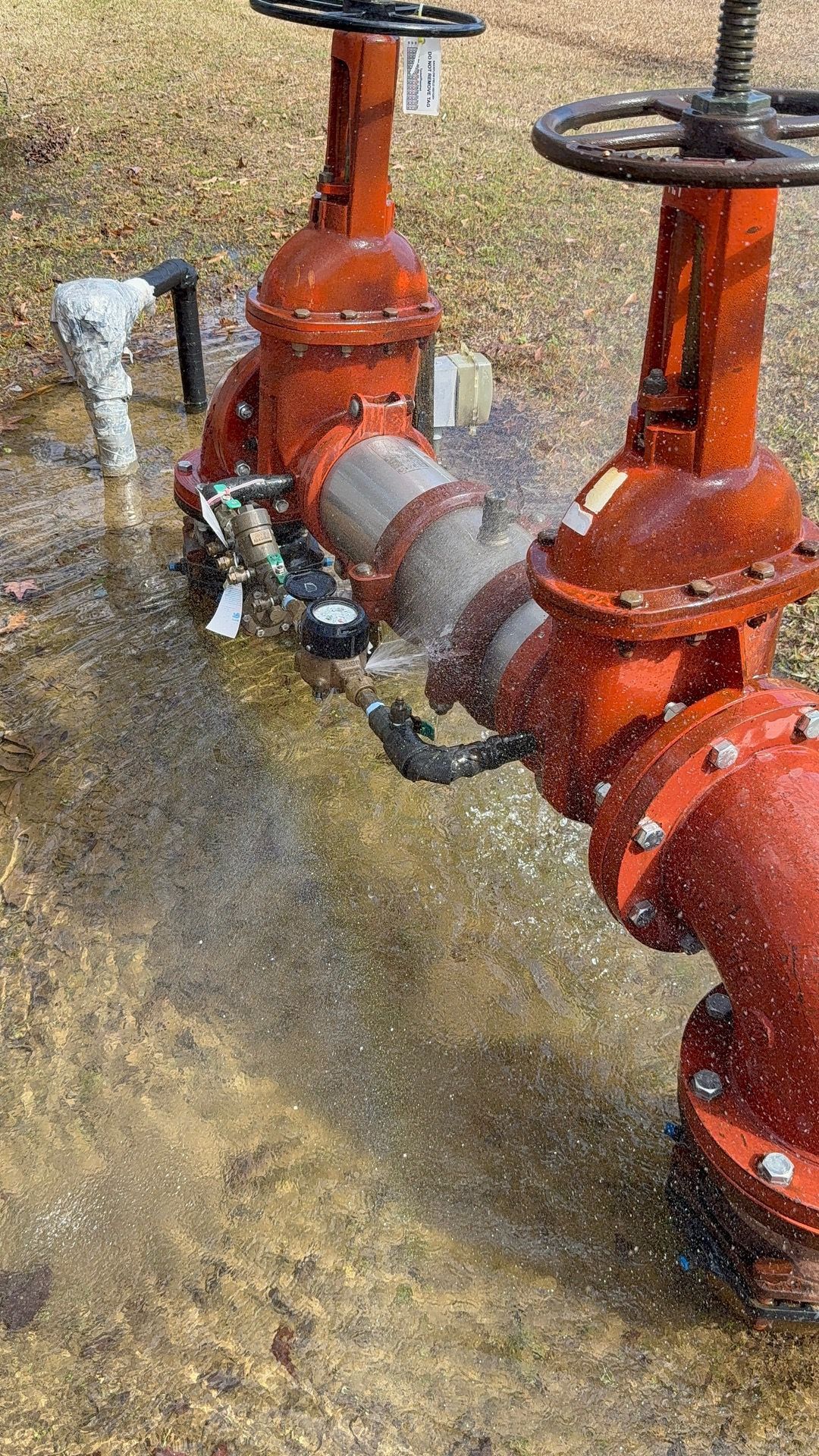 Rusty red industrial pipes and valves in muddy water near a grassy ground