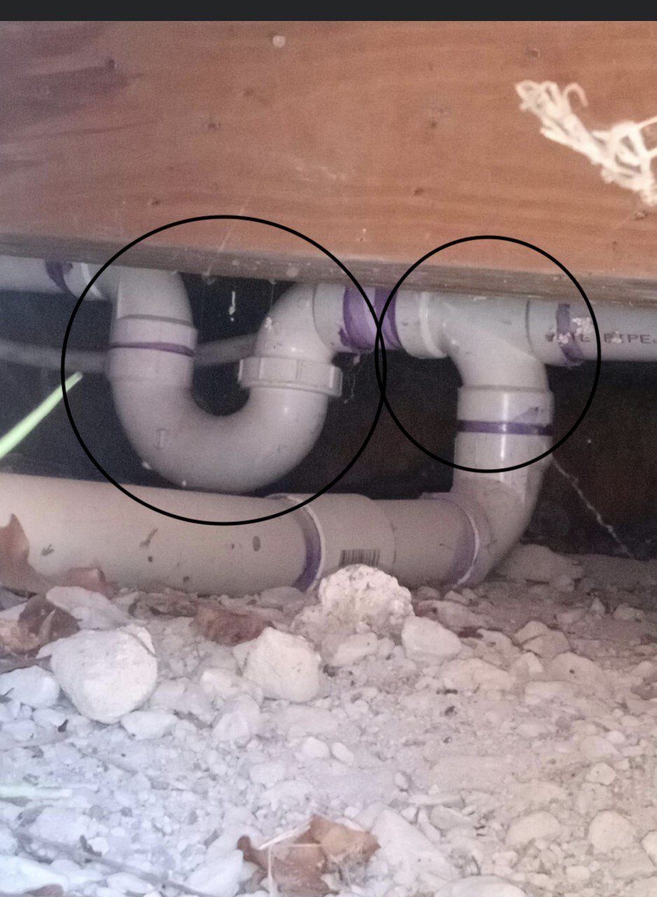 Two circled white PVC pipe traps under a wooden floor with insulation debris on the ground.