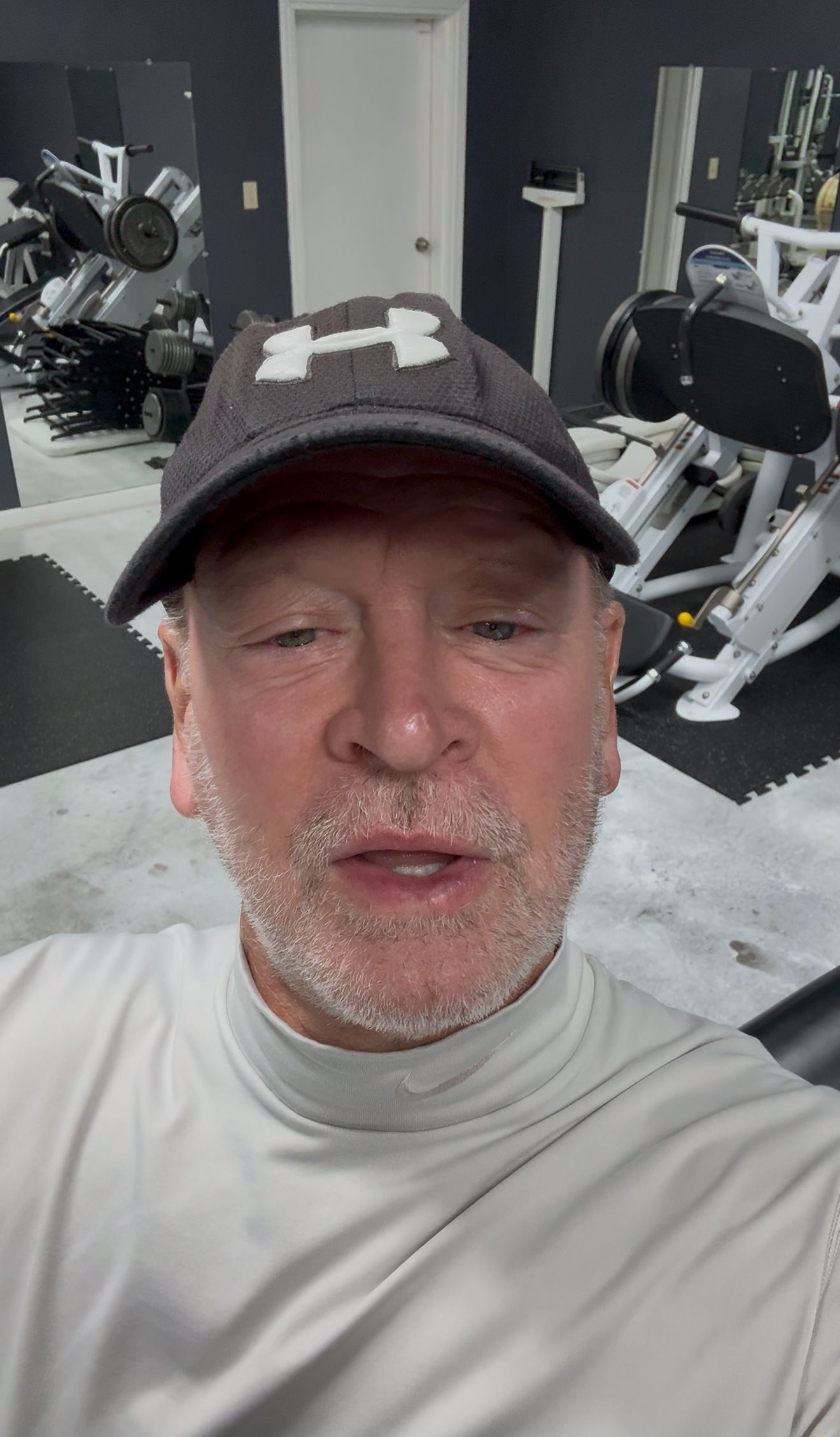Man in a gym wearing a cap and workout shirt, speaking toward the camera. Gym equipment in background.