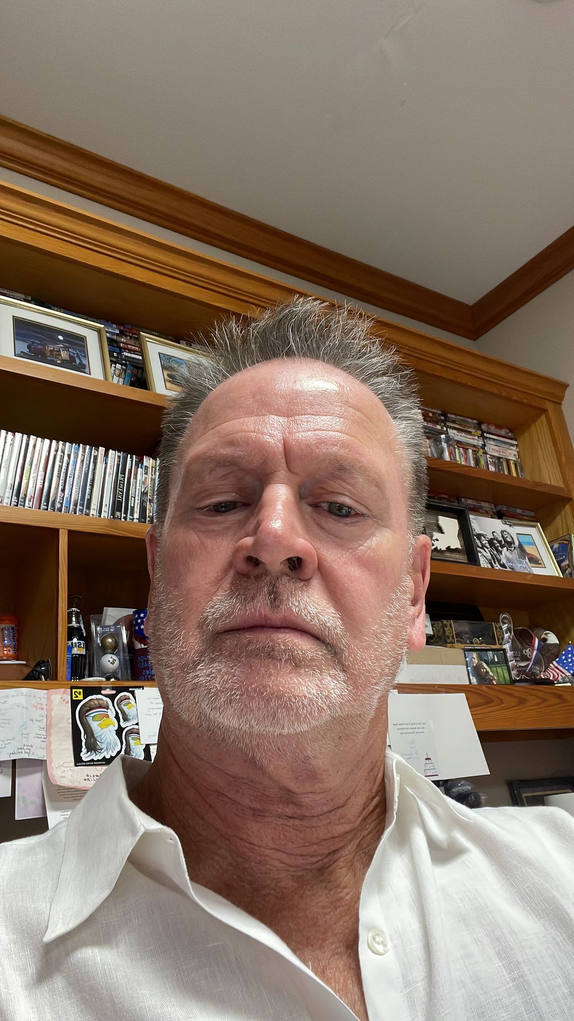 Man taking a selfie indoors in front of wooden shelves, wearing a white shirt