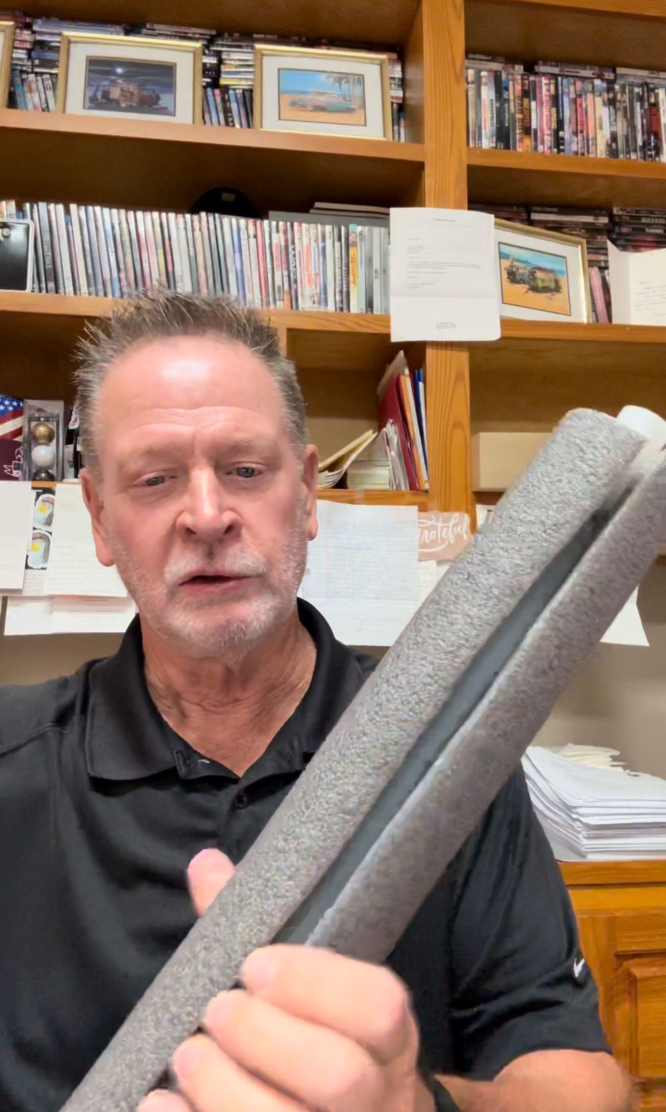 Man holding gray foam tube, indoors, speaking. Bookshelves in the background.