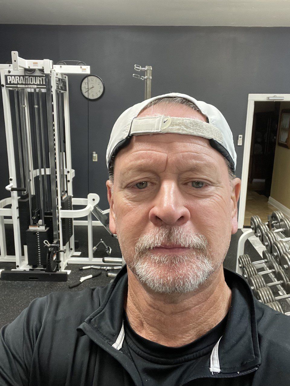 Man taking a selfie in a gym, wearing a backward cap and black shirt, with exercise equipment behind him