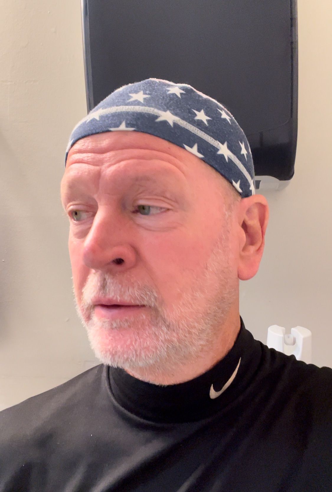 Man in a star-patterned bandana and black shirt, looking left against a plain indoor wall.