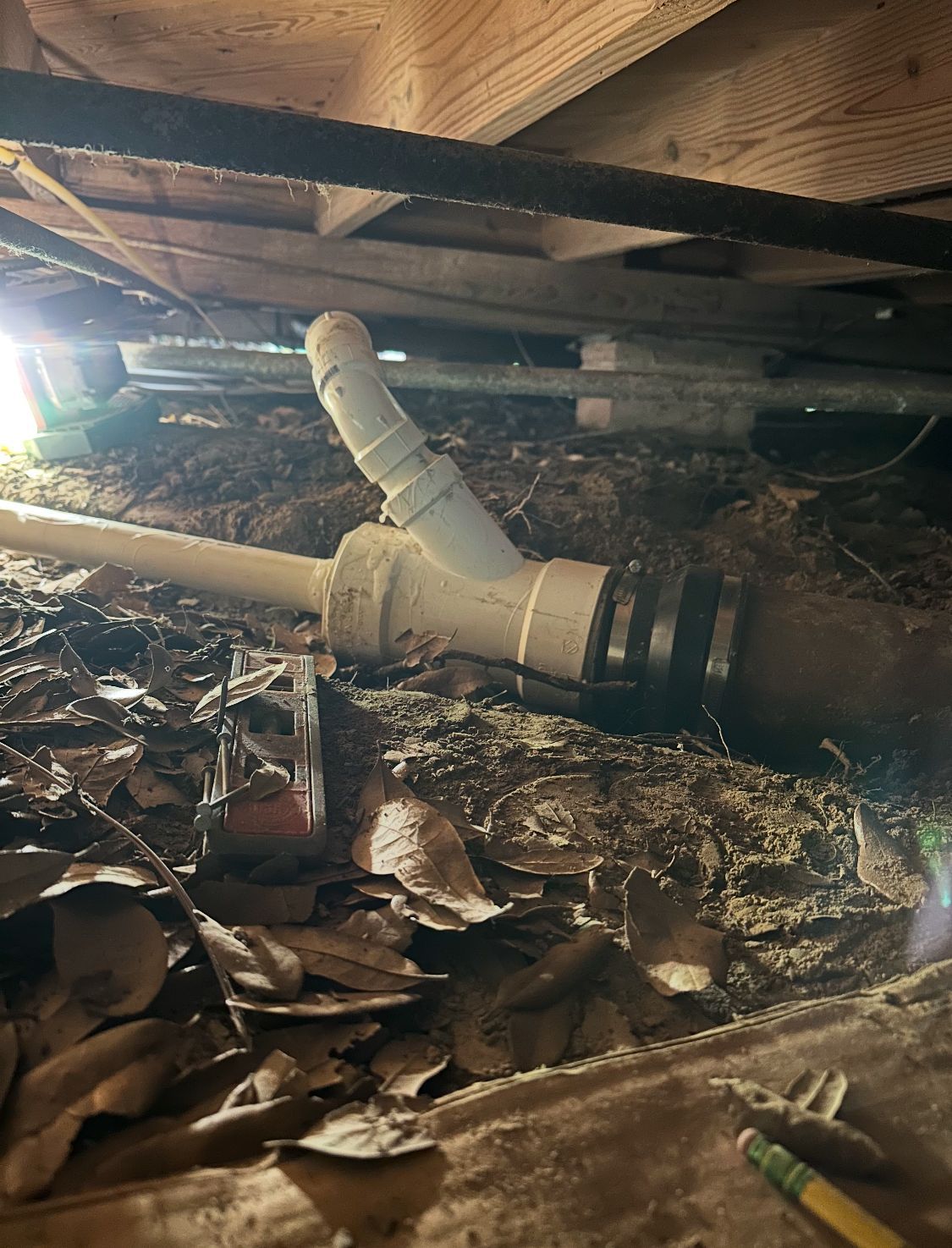 Underneath a house, plumbing pipes connect with a Y-shaped fitting and dark, decaying debris.