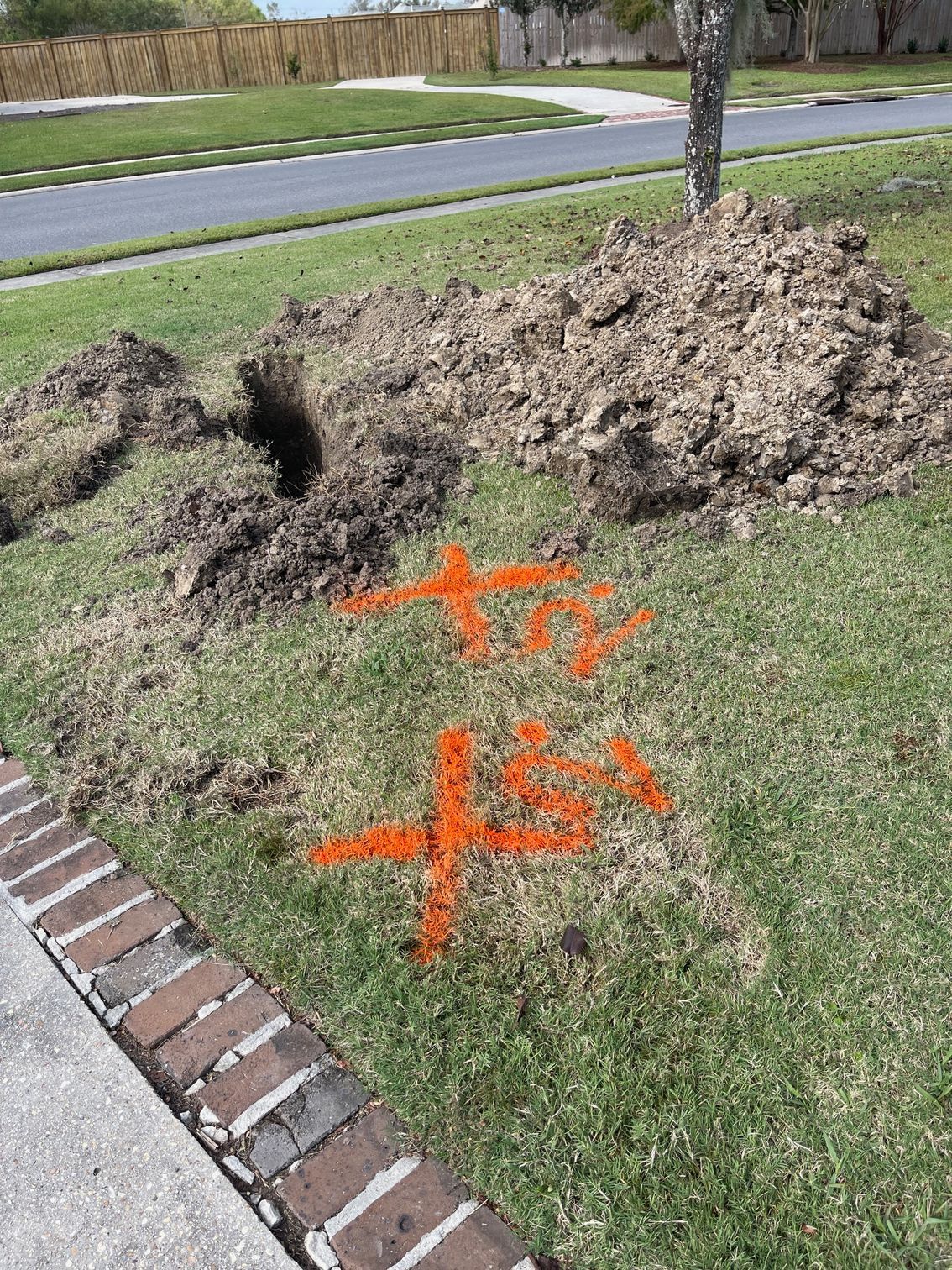 Lawn dug up near a sidewalk with orange spray paint markings and a pile of dirt.