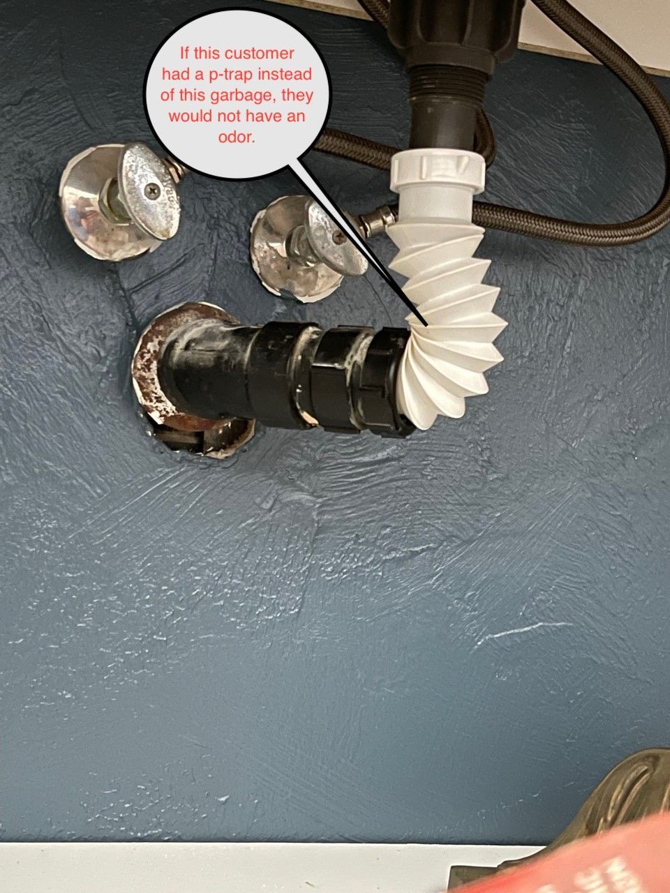 Plumbing under a sink: white flexible pipe, black drainpipe, silver faucet fixtures, blue wall.