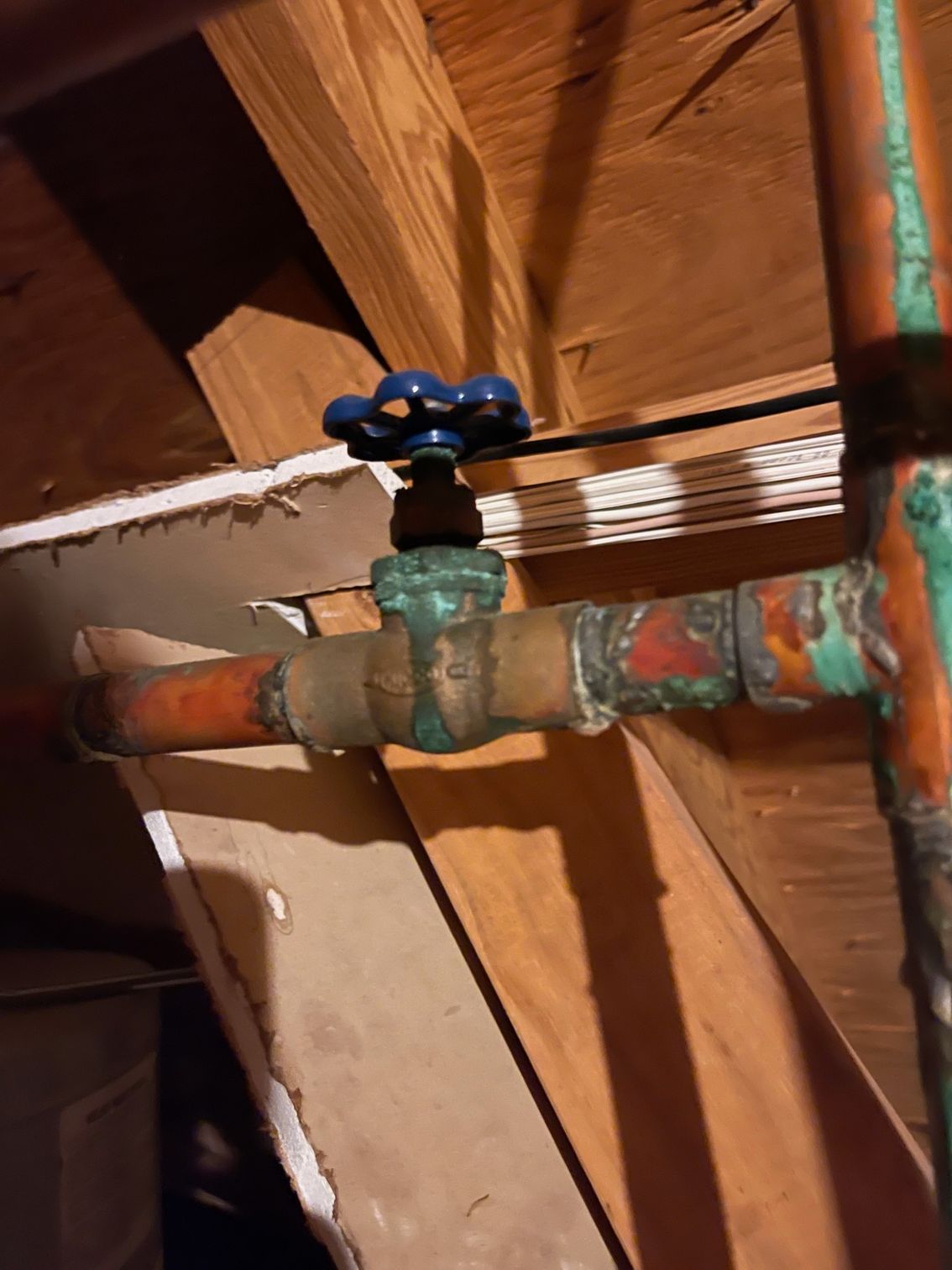 Old copper pipe with green valve under wooden ceiling, showing corrosion and a blue handle