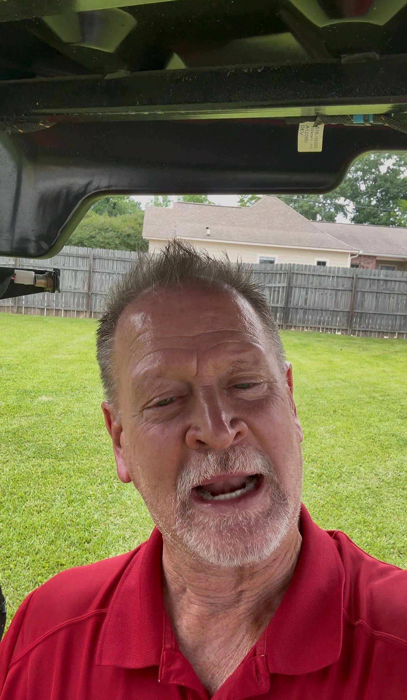 Man in a red polo shirt speaking inside a vehicle, with a grassy yard and fence behind him.