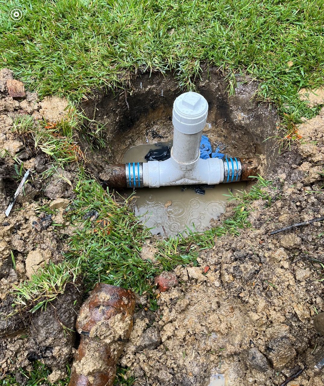 A shallow hole in grass containing a water pipe with a T-shaped connection and pooling water.