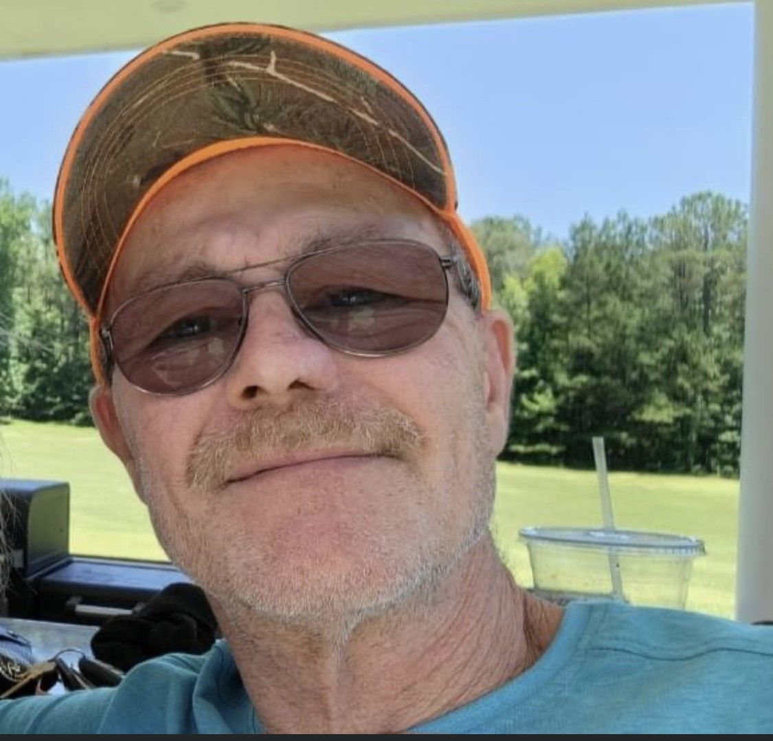 Man in sunglasses and a hunting hat smiles outdoors.