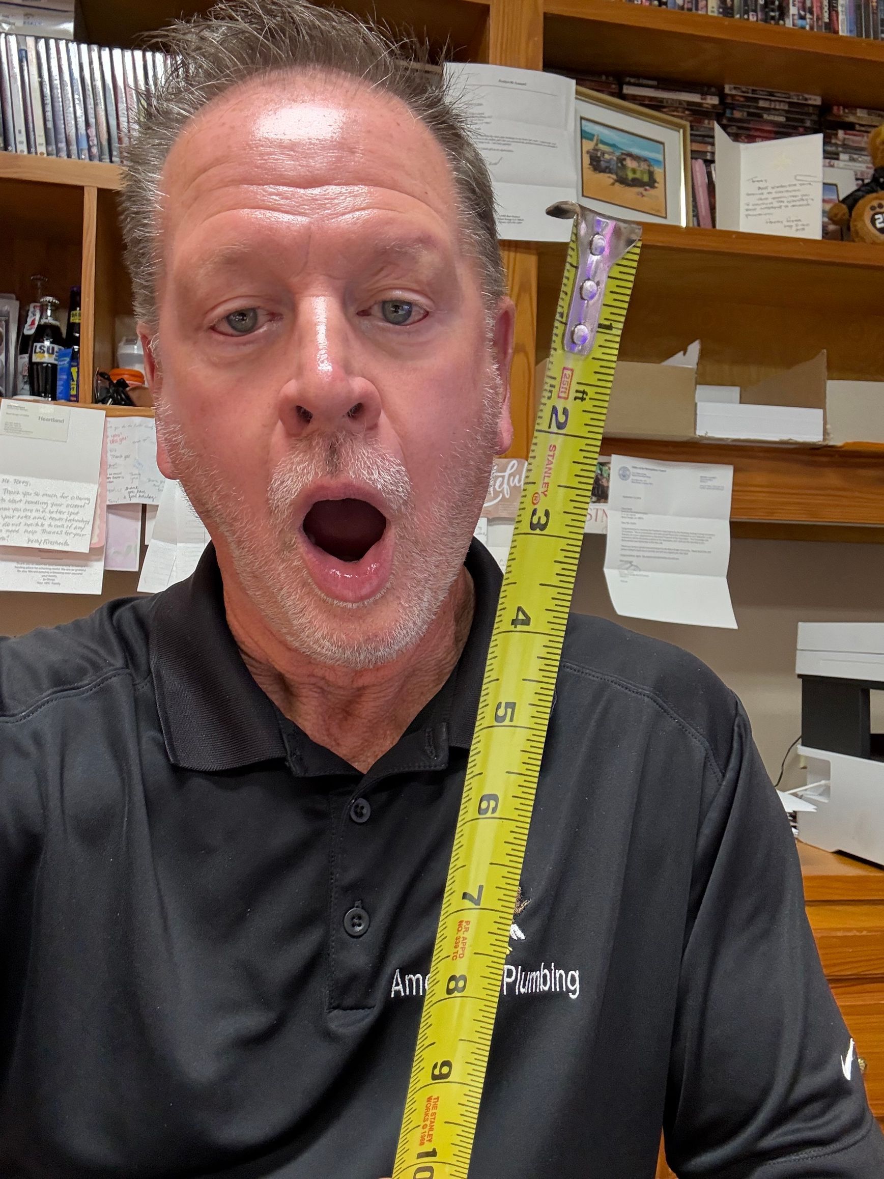 Man in black shirt in a storage room, holding a yellow measuring tape with mouth open in surprise.