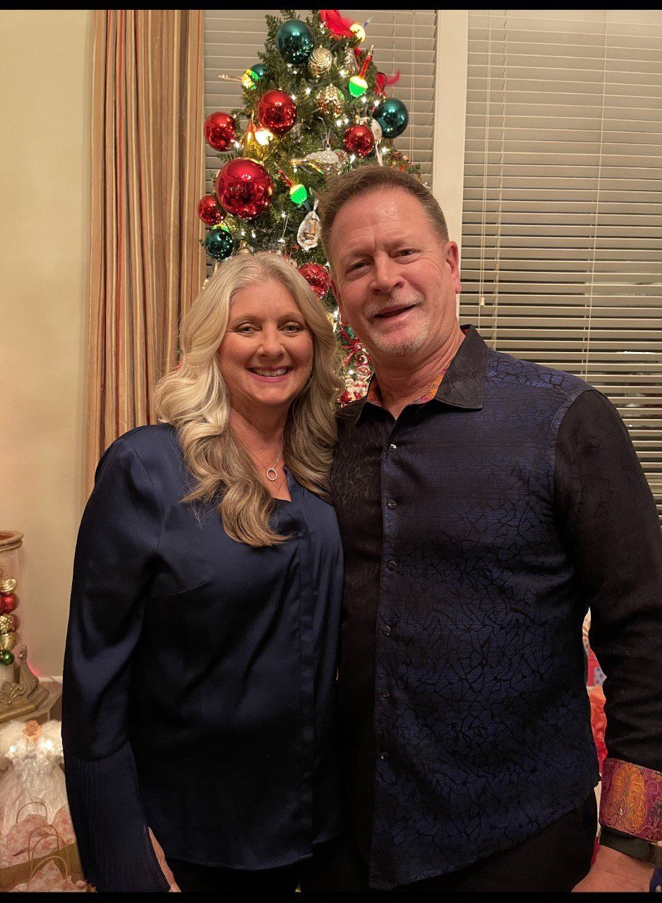 Couple smiling in front of a Christmas tree, the woman in blue, the man in a patterned shirt.
